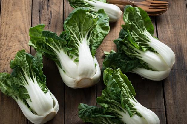 Five Pak Choi 'White' Baby Bok Choi plants, with their vibrant green leaves and crisp white stalks, rest elegantly on a wooden surface.