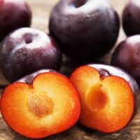 Several whole Prunus 'Spring Satin™ Plumcot' Dwarf 7" Pot plums with dark purple skin are shown, one cut in half to display its orange flesh and pit.
