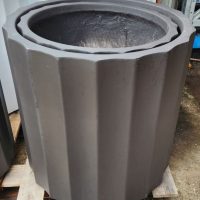 The GardenLite Grecian Cylinder Black S 39x39cm planter, featuring a dark matt finish and vertical ridges, is shown outdoors on a wooden pallet.