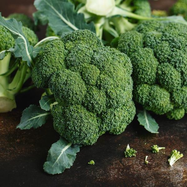 Fresh broccoli heads with leaves are displayed on a dark countertop, alongside the intricate, emerald-green spirals of Broccoli 'Romanesco' (Copy).