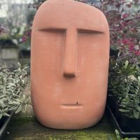 A Moai Statue Terracotta 30x29x46cm stands amidst potted plants displaying various shades of green and purple.