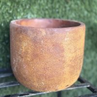 A large, round Portica Egg Pot Rust M 16x13cm with a rough texture sits on a black metal stand against a background of green artificial grass, making it an ideal garden planter.