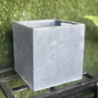 A plain, gray, square concrete planter sits on a metal rack with a backdrop of green foliage, reminiscent of the sleek design of the Portica Egg Pot Rust M 16x13cm (Copy).