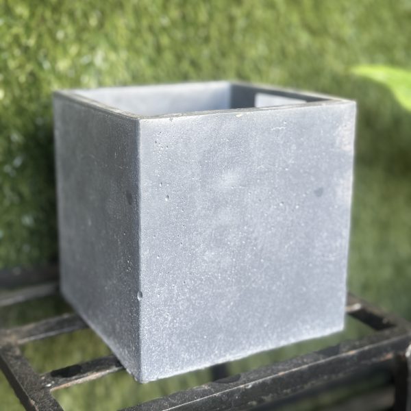 A plain, gray, square concrete planter sits on a metal rack with a backdrop of green foliage, reminiscent of the sleek design of the Portica Egg Pot Rust M 16x13cm (Copy).