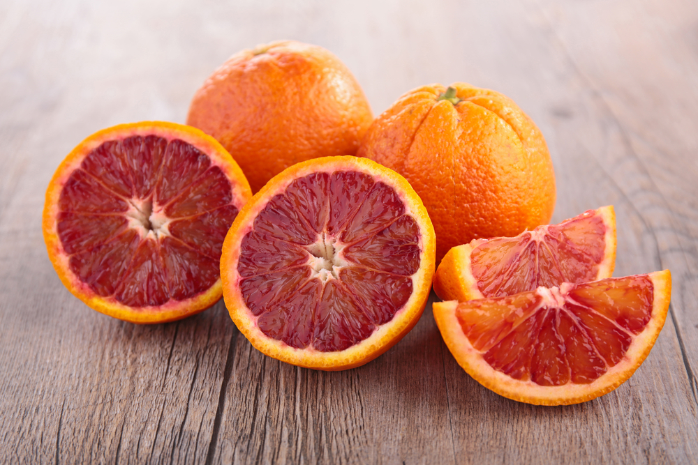 Two whole and two halved blood oranges, displaying vibrant red flesh, rest on a wooden surface. These fruits highlight the rich hue and succulent nature of the prized Citrus Orange Tree 'Blood Orange' Dwarf from a 10" pot.