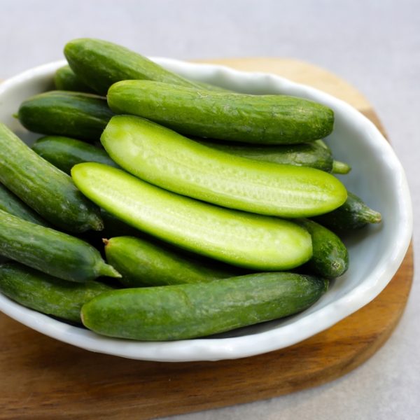A white bowl filled with whole and halved fresh mini cucumbers, including the 'Bebe Lebanese' variety from a 4" pot, on a wooden surface.