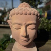 Close-up of the Terracotta Buddha Head 26x40cm sculpture, featuring a peacefully closed-eyed expression reminiscent of a Moai statue, set outdoors with surrounding greenery slightly out of focus.