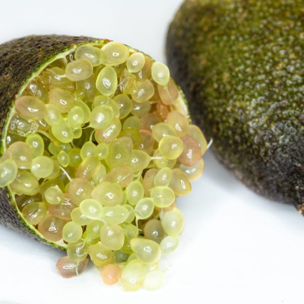 A Citrus 'Gold Pearl' Finger Lime from the "7" Pot" collection sliced in half, with its green and pinkish pulp spilling out like tiny gold pearls, rests on a white surface beside another intact citrus gem.