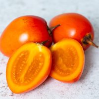 A trio of Solanum 'Denmark Gold' tamarillos, sourced from the Solanum family, are displayed on a textured surface; with two intact and one cut open to showcase its vibrant orange interior.