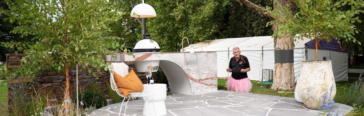 An outdoor stone patio features a pizza oven, table, and chair. A person stands nearby in casual attire with a pink tutu. Amidst the trees and a large tent, this setting perfectly complements the many benefits of gardening that surround it.