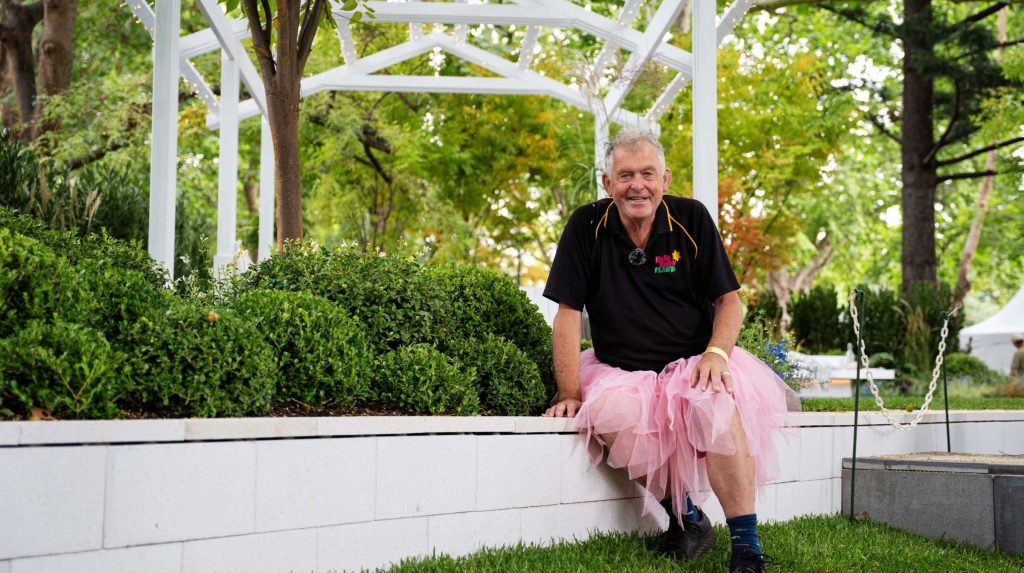 An older man enjoys the many benefits of gardening as he sits on a white ledge, wearing a black shirt and pink tutu. Behind him, trees sway gently near a charming white gazebo, offering a peaceful retreat in nature's embrace.