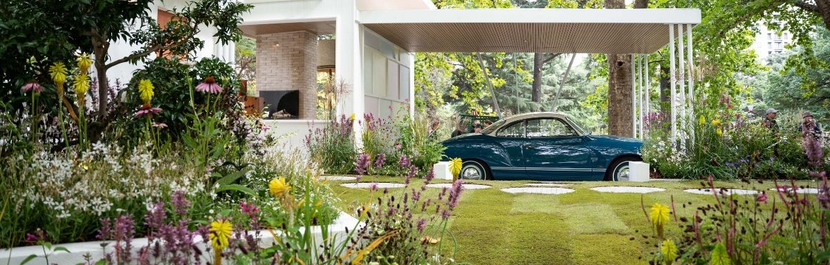A blue vintage car is parked under a modern white canopy, surrounded by a lush garden that highlights the many benefits of gardening with its colorful flowers and vibrant greenery.