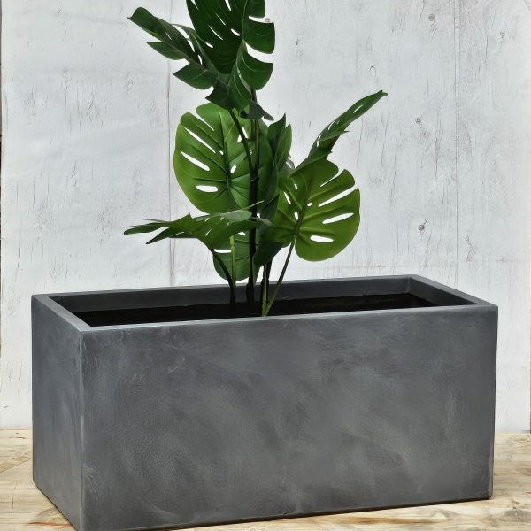 A tall potted plant with large green leaves in a rectangular gray planter, placed on a wooden surface against a textured light background.