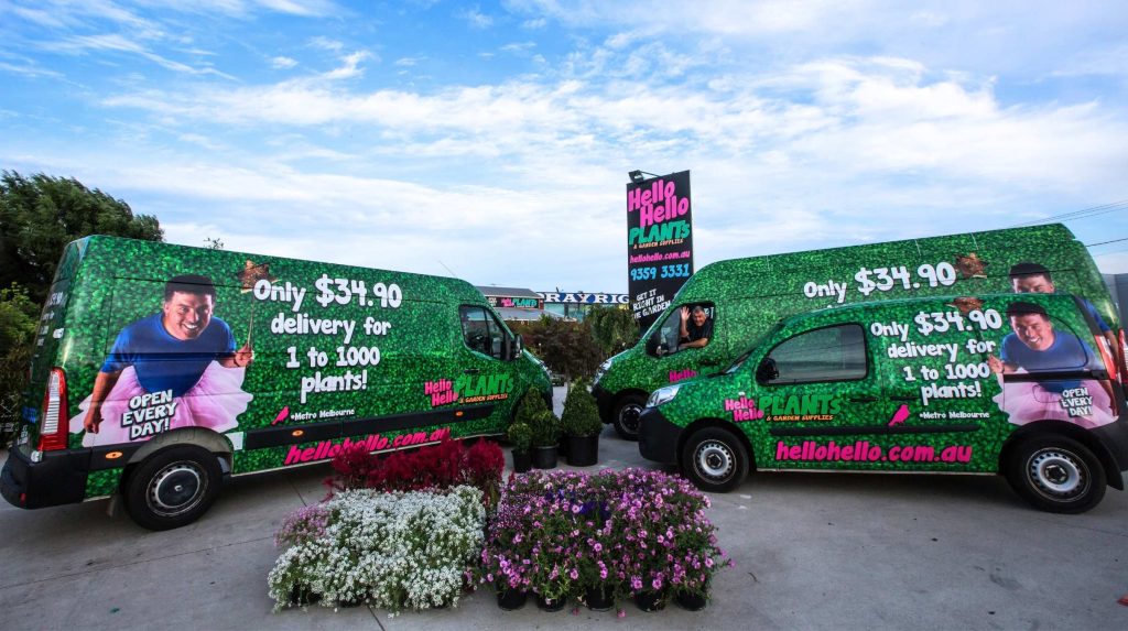 Two green vans with "Hello Hello Plants" advertising, pricing, and hiring information are parked near a display of various potted plants and flowers outside the store.