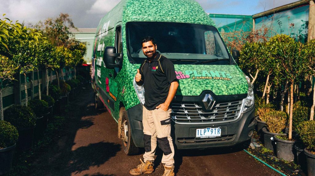 A man stands smiling next to a green delivery van with "Hello Plants & Garden Supplies" written on it, surrounded by potted plants and trees outdoors, with a sign in the window that reads "Now Hiring.