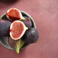 A bowl featuring a mix of whole and halved Ficus 'Violette De Bordeaux' figs sits on a pink surface.