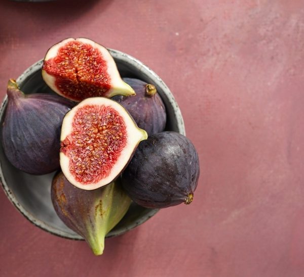 A bowl featuring a mix of whole and halved Ficus 'Violette De Bordeaux' figs sits on a pink surface.