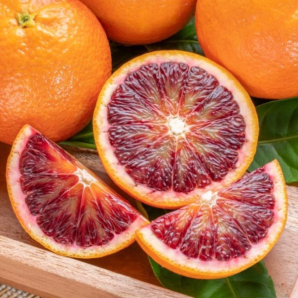 A wooden tray holds whole and sliced Citrus ‘Arnold’ Blood Orange 4L, showing vibrant red flesh, surrounded by fresh green leaves.