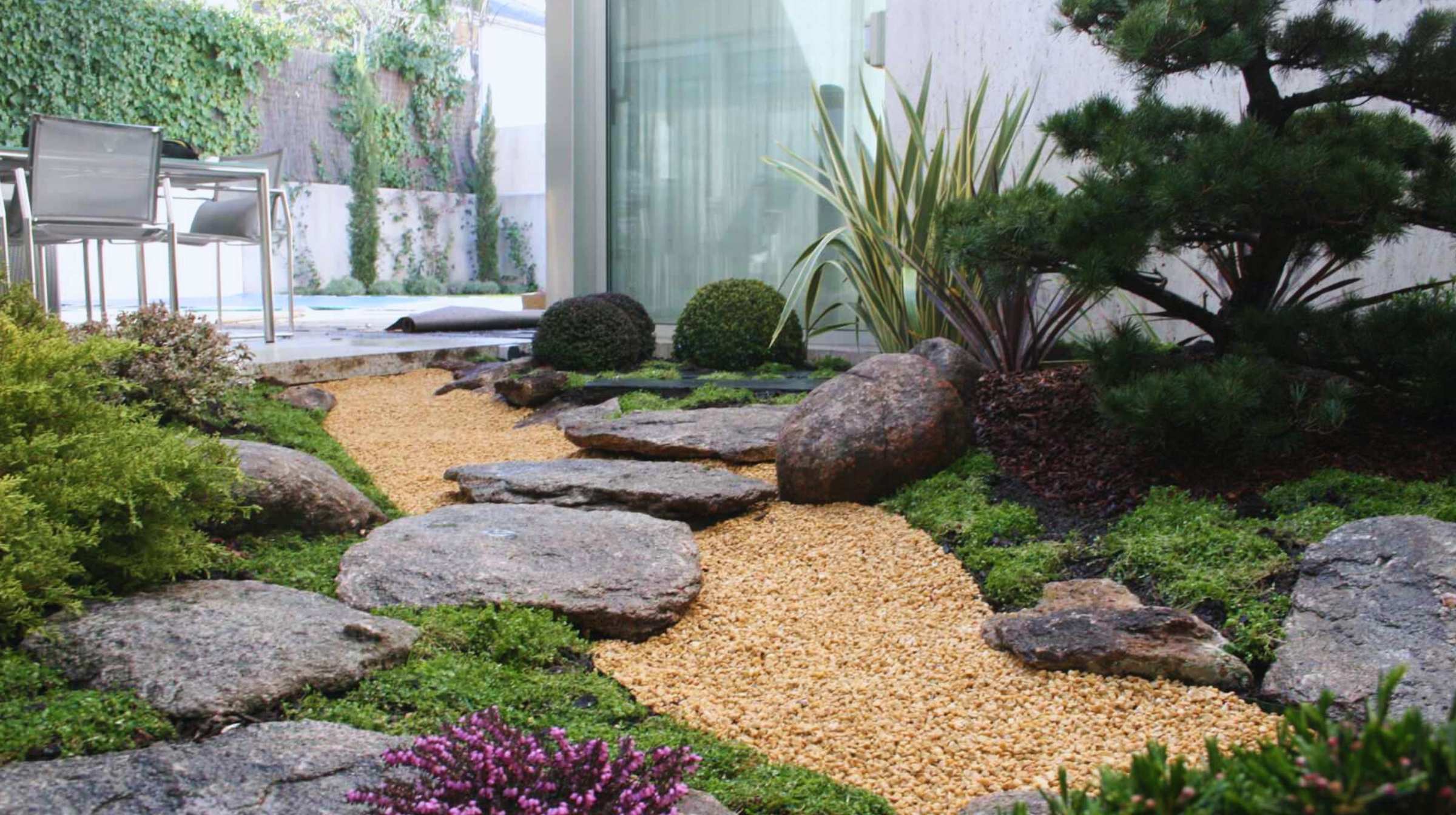 A landscaped garden with large stepping stones and a gravel path, surrounded by green shrubs and bushes, captures the beauty of Japanese gardens. A patio with chairs sits in the background, inviting relaxation.