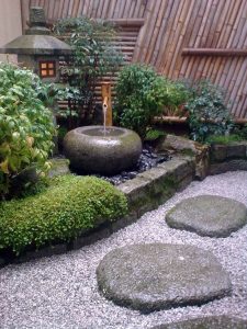 A Japanese garden showcasing the beauty of Japanese gardens with a stone water basin, bamboo spout, moss-covered stones, gravel path, lush green plants, and a bamboo fence in the background.