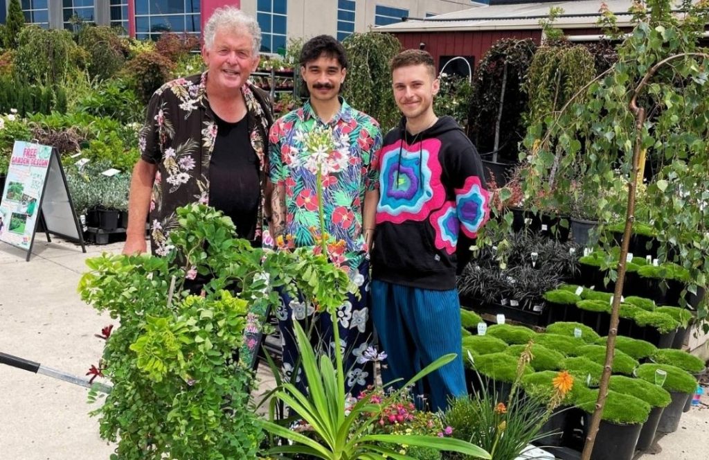 Three people stand together outdoors at a plant nursery, surrounded by various green plants and shrubs, ready to get the right plants for their garden, with buildings visible in the background.