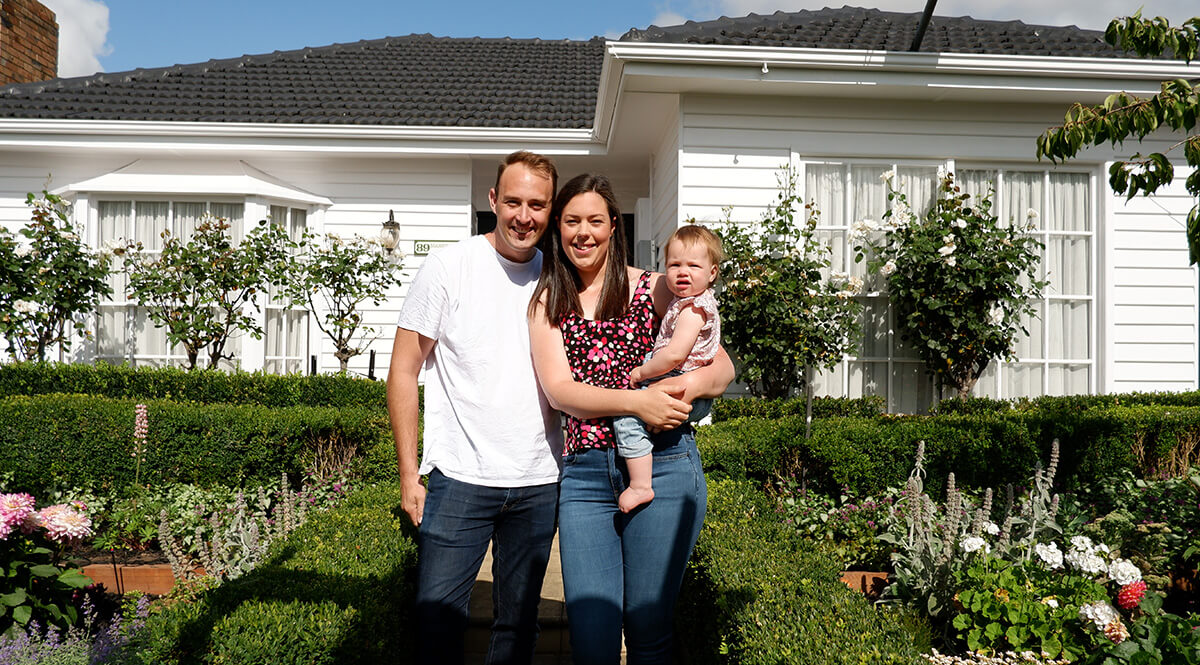 A man, woman, and baby stand together in front of a white house with a garden and hedges on a sunny day, showcasing how curb appeal can increase the value of your property.