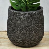 A large, textured dark gray planter with a green monstera plant, placed on a wooden surface against a light-colored wall.