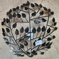 The Raja TeaLight Wall Art Black 46x10cm is a round metal wall decor with a symmetrical tree branch pattern and black leaves, displayed on a concrete surface. A white barcode sticker is near the bottom center.