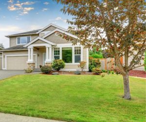 Two-story suburban house with gray siding, a covered front porch, and attached garage—set behind a well-kept lawn and tree—perfect features to increase the value of your property.
