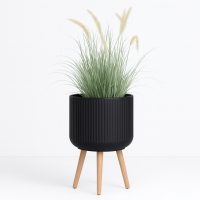 The GardenLite Corrugated Cylinder with Legs Black M 39x38cm planter holds tall ornamental grass with thin green leaves and feathery beige plumes, displayed against a white background.