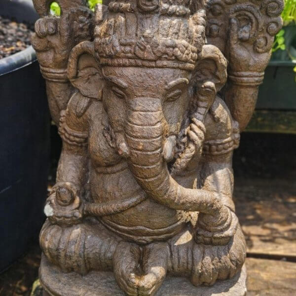 A stone statue of Ganesha, the Hindu deity with an elephant head, seated with one hand raised and detailed carvings, surrounded by greenery.