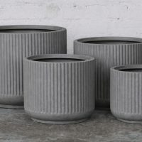 Four grey, ribbed, cylindrical planters of varying sizes arranged on a concrete floor against a white brick wall.